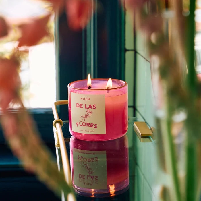 A pink jar candle with multiple wicks burning inside, placed on a surface with flowers in the background.