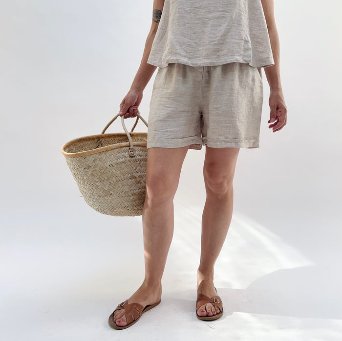 Person wearing a beige linen tank top and matching linen shorts holding a woven basket on a white background.