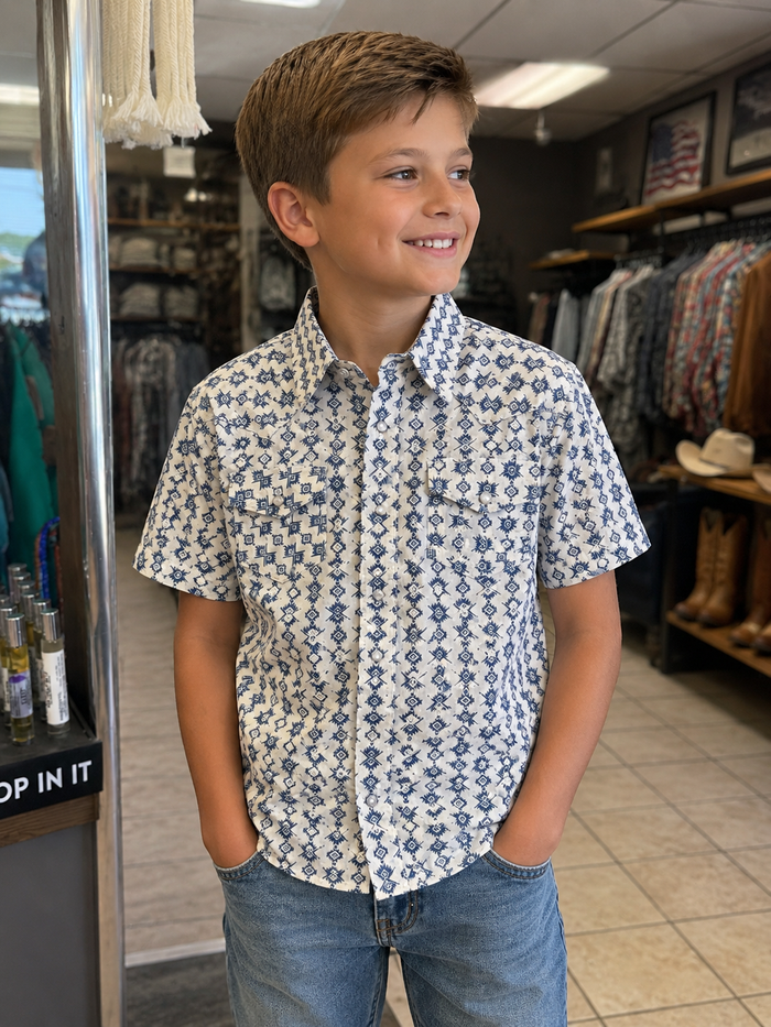A young boy smiling in a clothing store, wearing a short-sleeve button-down shirt with a blue and white geometric pattern and blue jeans.