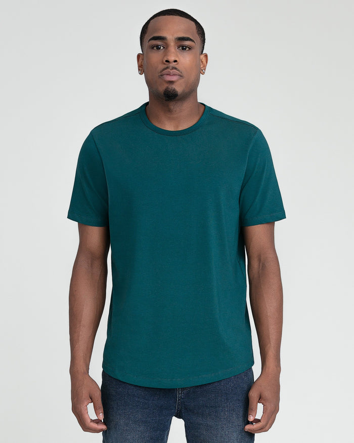 Man wearing teal crew neck short-sleeve t-shirt with dark jeans against neutral background.