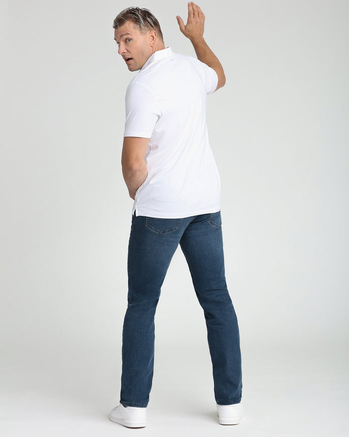 Man wearing white short-sleeve polo shirt with dark blue jeans and white sneakers, shown from back at an angle.