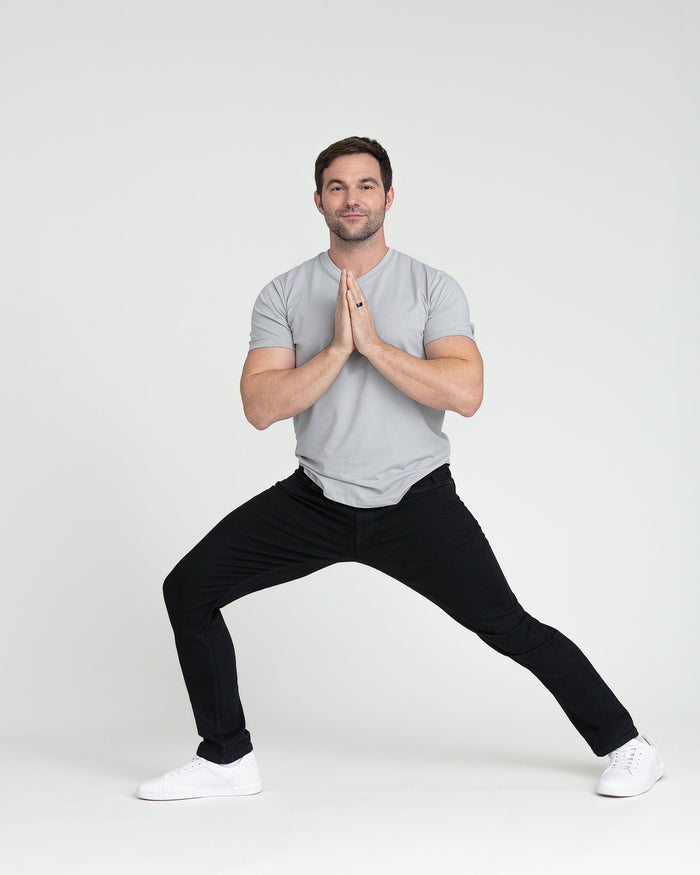 Man wearing gray t-shirt and black slim fit pants in a wide-legged stance with hands pressed together.