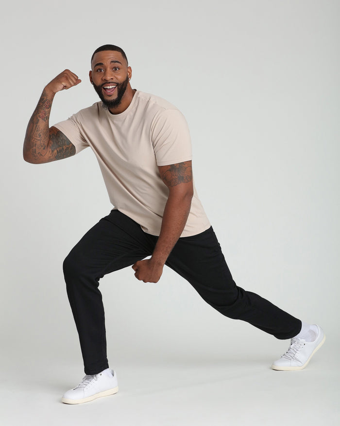 Man wearing beige short-sleeve t-shirt and black pants with white sneakers, posed in dynamic stance.