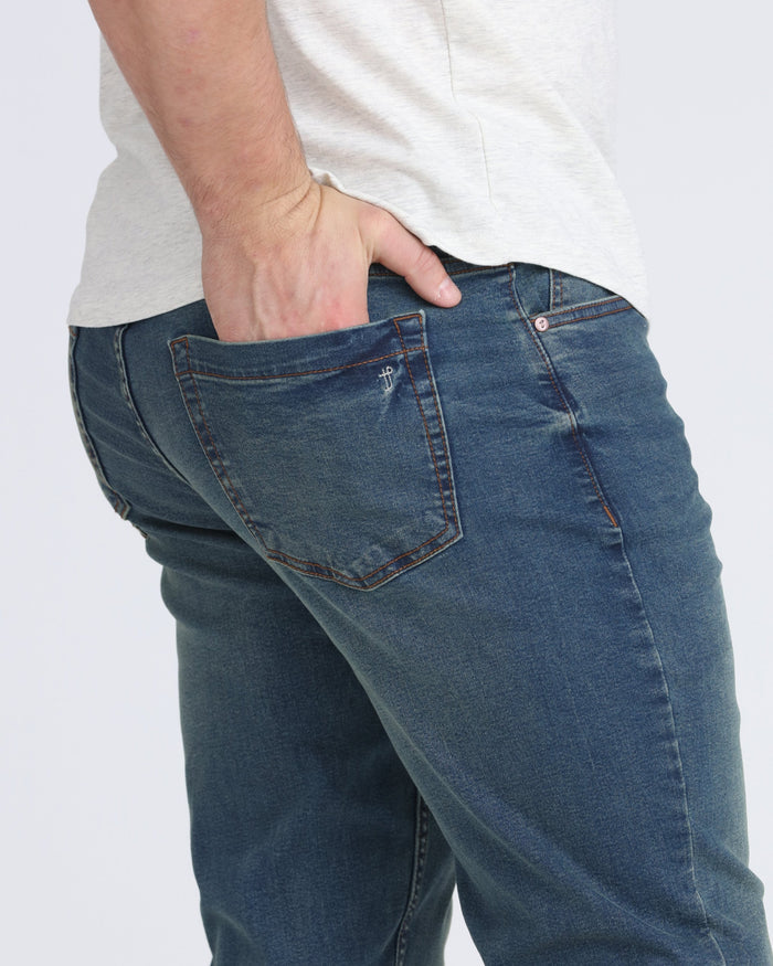Man wearing medium-wash denim jeans with relaxed thigh fit and brown contrast stitching, paired with cream t-shirt.