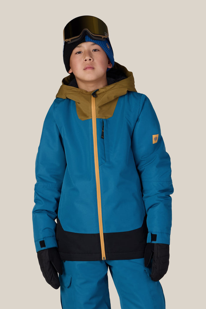 A child wearing the 686 Boys’ Gateway™ Insulated Jacket by 686, along with blue pants, black gloves, a blue beanie, and ski goggles, stands against a plain light background and looks at the camera.
