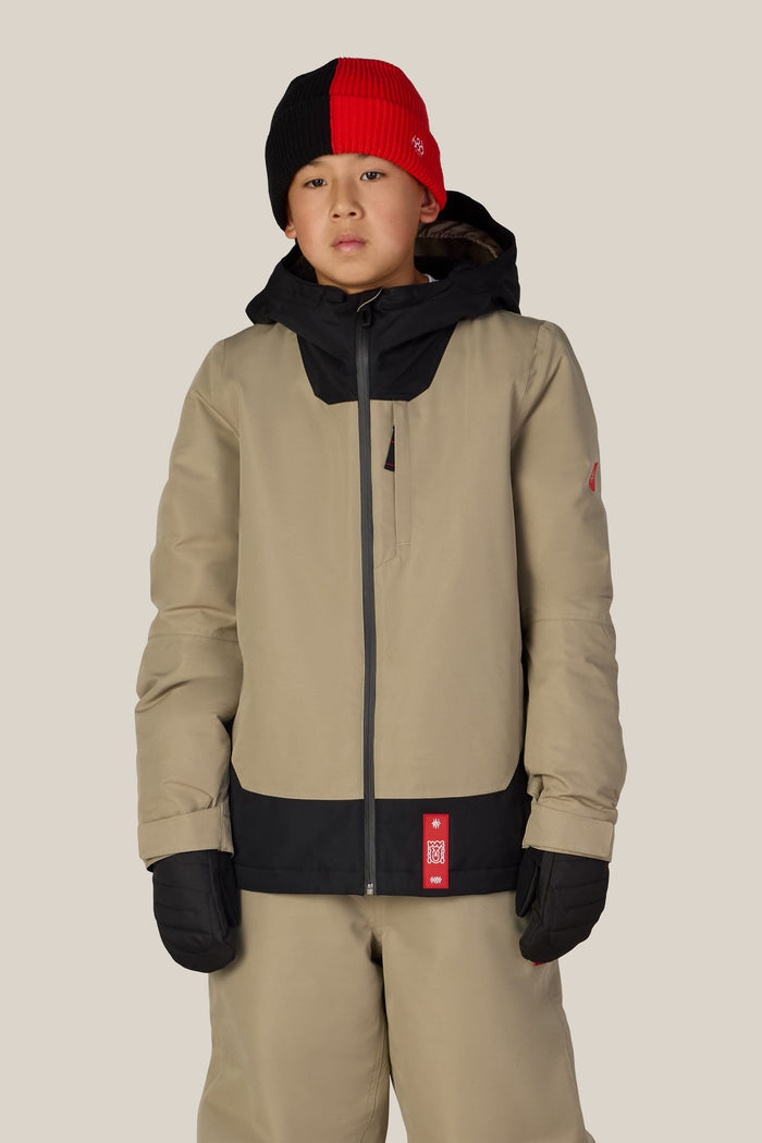 A child wearing the 686 Boys’ Gateway™ Insulated Jacket by 686, paired with tan pants, black gloves, and a red and black knit beanie, stands against a plain light background.