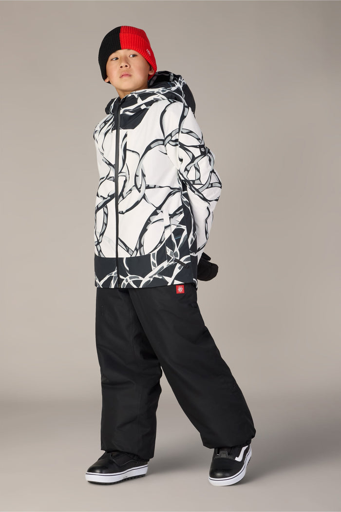 A child stands wearing the 686 Boys’ Gateway™ Insulated Jacket by 686, paired with black snow pants, black and white sneakers, and a red and black beanie, looking to the side with hands behind their back.