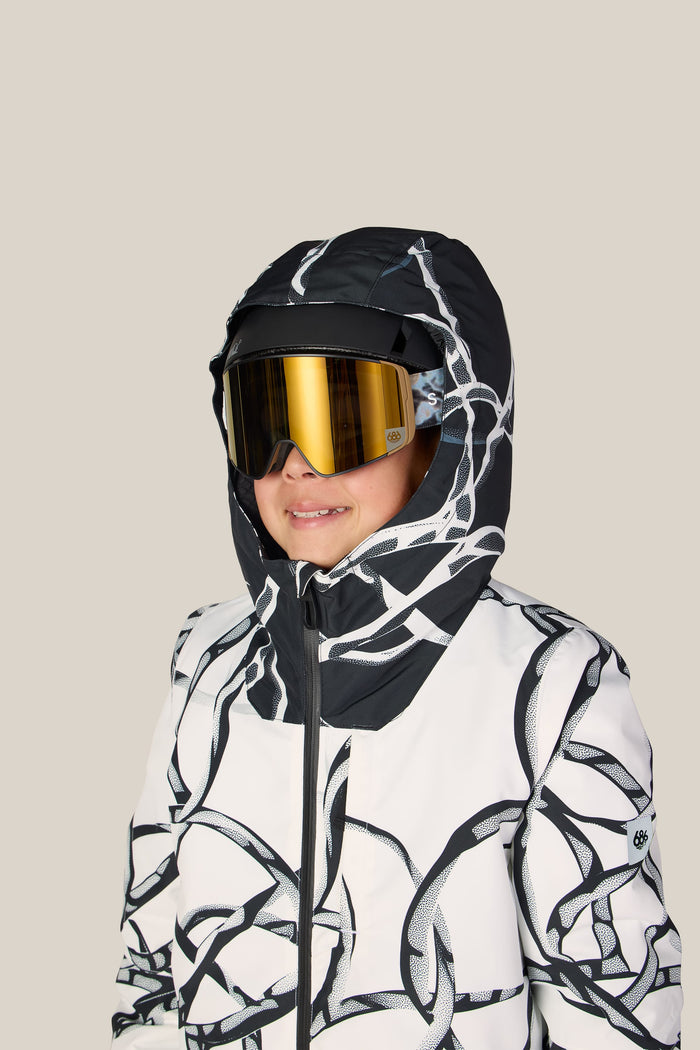A child wearing the 686 Boys’ Gateway™ Insulated Jacket by 686, featuring a hooded black and white design with abstract patterns, along with gold ski goggles, is smiling and facing slightly to the right against a plain light background.