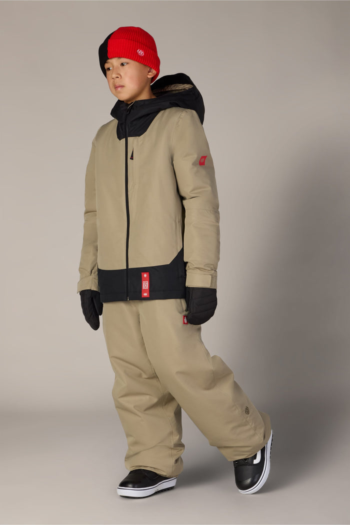 A child wearing the 686 Boys’ Gateway™ Insulated Jacket by 686, tan winter pants, black gloves, black shoes, and a red and black beanie stands against a plain background.