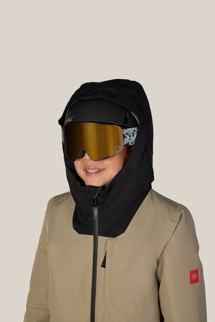 A child smiles against a plain beige background while wearing the 686 Boys’ Gateway™ Insulated Jacket by 686, featuring a tan color with a red patch, black hood, and reflective ski goggles.