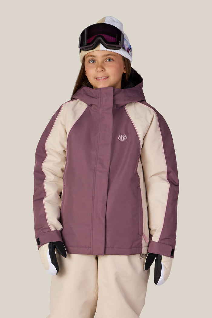 A young person in the 686 Girls’ Hologram™ Insulated Jacket by 686, paired with cream ski pants, white gloves, and ski goggles, smiles at the camera against a plain, light background.