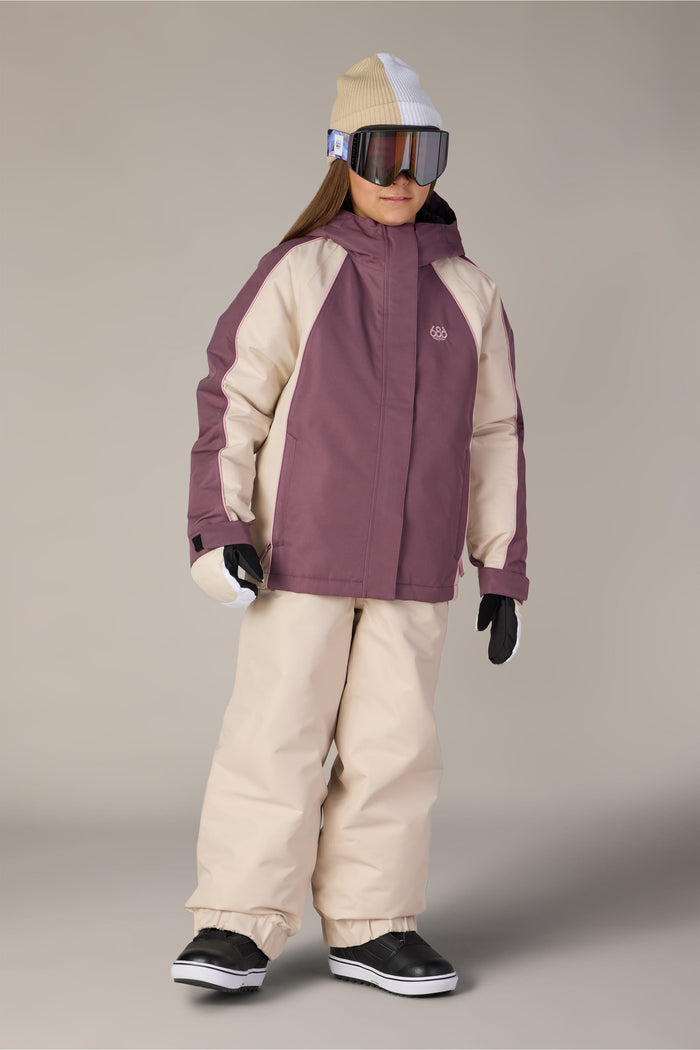 A child wearing the 686 Girls’ Hologram™ Insulated Jacket in purple and beige, paired with matching waterproof pants, a beige and white beanie, large ski goggles, black gloves, and black snow boots, standing against a plain background.
