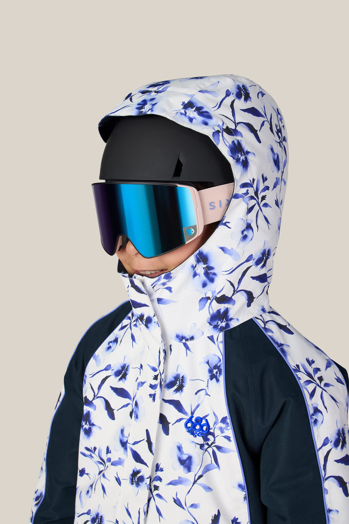 A person is wearing the 686 Girls’ Hologram™ Insulated Jacket by 686, featuring a blue floral pattern and hood, paired with a black ski helmet and reflective ski goggles against a plain beige background.