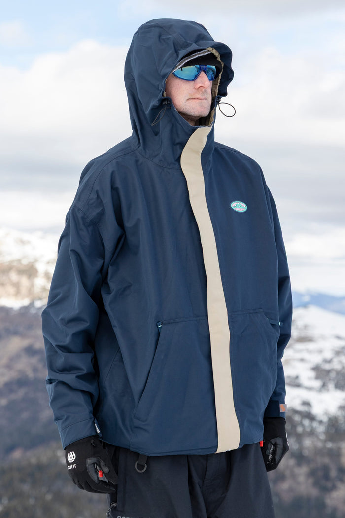 A person wearing the 686 Men’s 1994 Liberty Jacket by 686, along with blue sunglasses and black gloves, stands outdoors against a backdrop of snowy mountains and a cloudy sky.