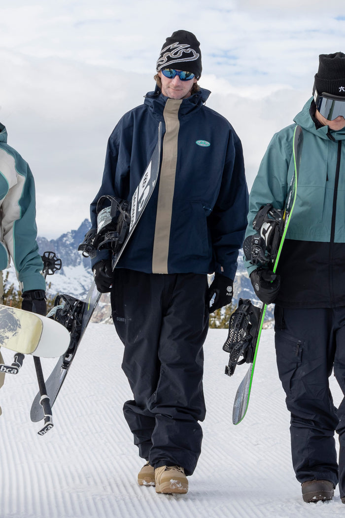 Three people in winter gear walk on snow, carrying snowboards. The central person is wearing the 686 Men’s 1994 Liberty Jacket, a beanie, blue sunglasses, and tan boots. Snowy mountains and a cloudy sky are visible in the background.