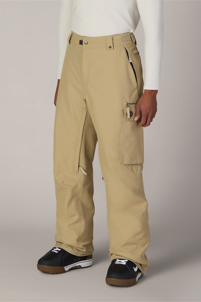 A person wearing the 686 Men’s 686 Nightline Cargo Pant in tan, paired with a white long-sleeve shirt and black snow boots, stands against a plain background.