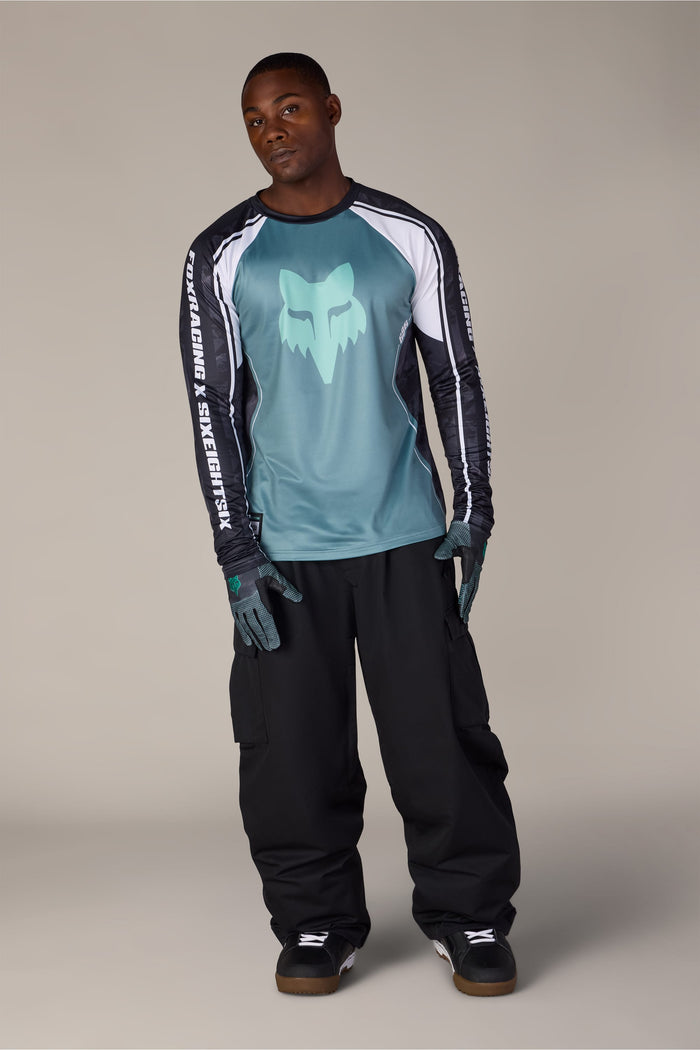 A man models a 686 Men’s Fox Racing Base Layer LS with a teal logo, black cargo pants, and gloves.