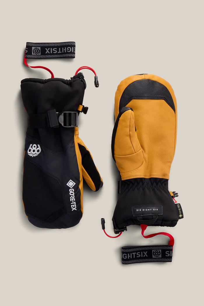 Two 686 Men’s GORE-TEX® Leather Linear™ Mitts, one in black and the other in tan with black accents, are displayed side by side on a beige background. Both mitts feature wrist straps labeled “686,” and the black mitten displays a GORE-TEX® logo.