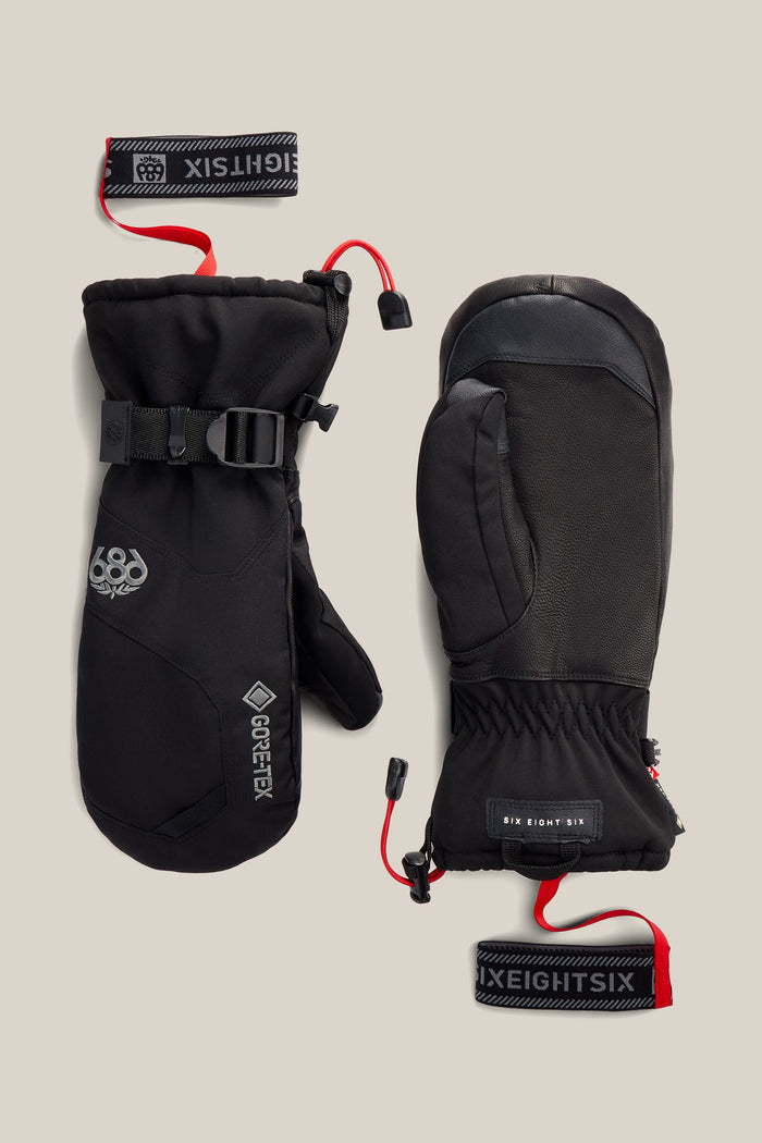 A pair of 686 Men’s GORE-TEX® Leather Linear™ Mitts in black, featuring red wrist straps, adjustable buckles, and visible 686 branding. One mitten displays the 686 logo and a GORE-TEX label, while the other features additional branding and textured palm detailing.