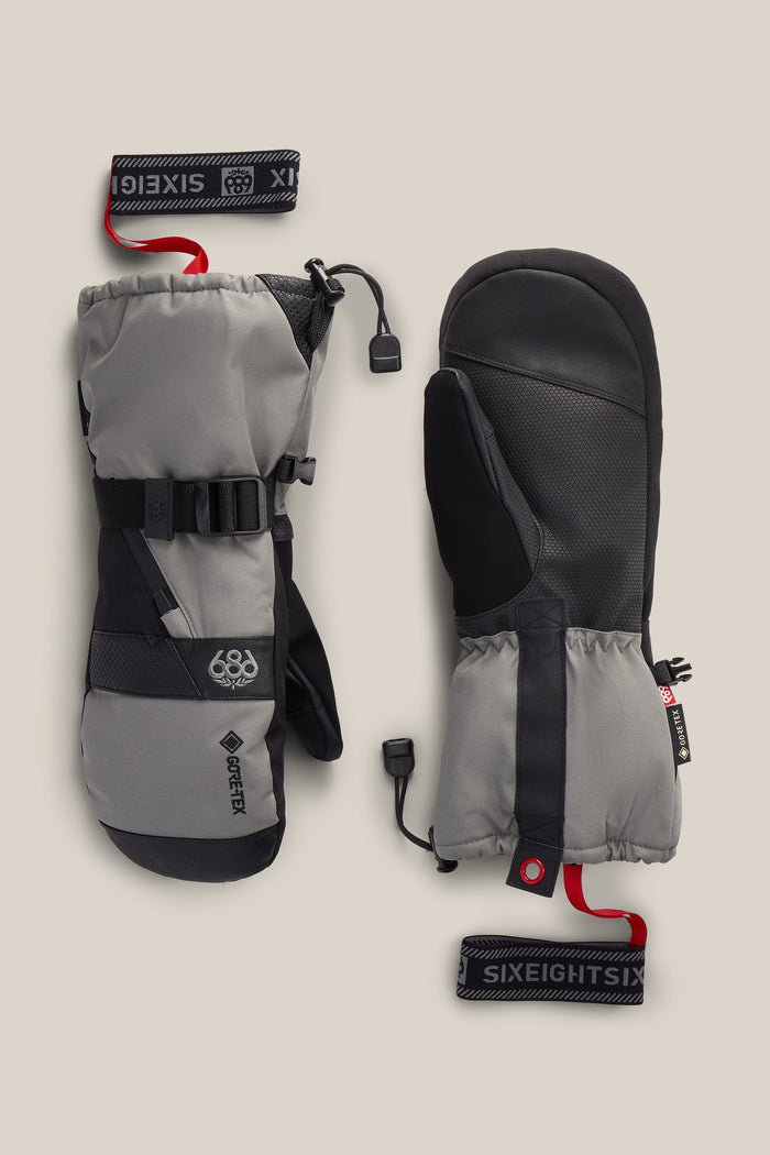 Two gray and black 686 Men’s GORE-TEX® SMARTY® 3-In-1 Gauntlet Mitts with adjustable straps and red 686 wrist leashes, displayed on a beige background. One mitten displays the palm side, while the other shows the back.