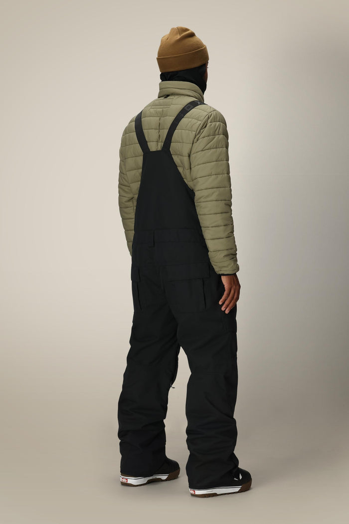 A person stands facing away, wearing a brown beanie, olive puffer jacket, and the 686 Men’s Hot Lap™ Insulated Bib in black with black shoes, against a plain light background.