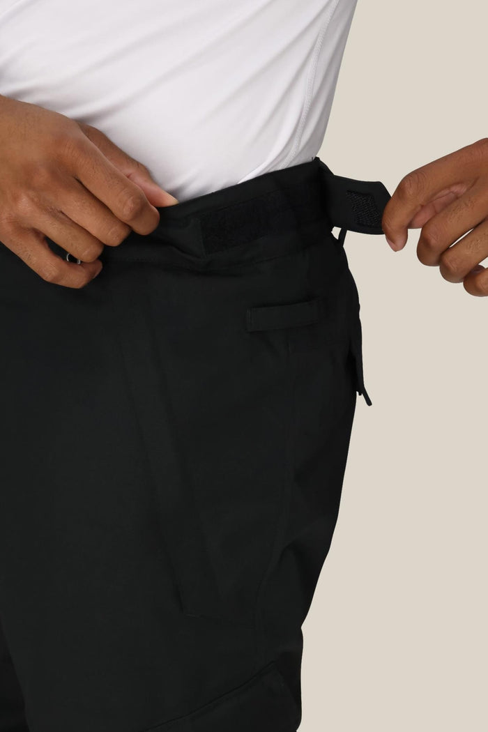 A person adjusts the waistband of the 686 Men’s Infinity™ Insulated Cargo Pant using a Velcro strap. Only their hands, the waistband, and part of their white shirt are visible against a light background.