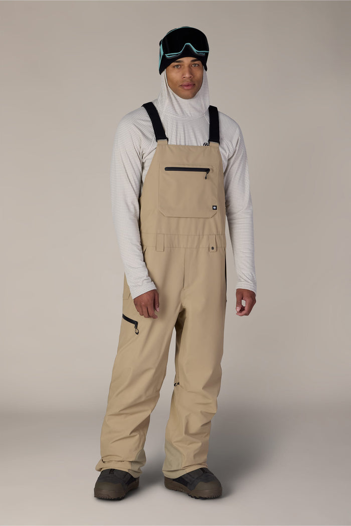 A person stands against a plain background wearing the 686 Men’s Lockup™ Shell Bib in beige by 686, paired with a white hooded top, black gloves, snow boots, and black ski goggles resting on their head.