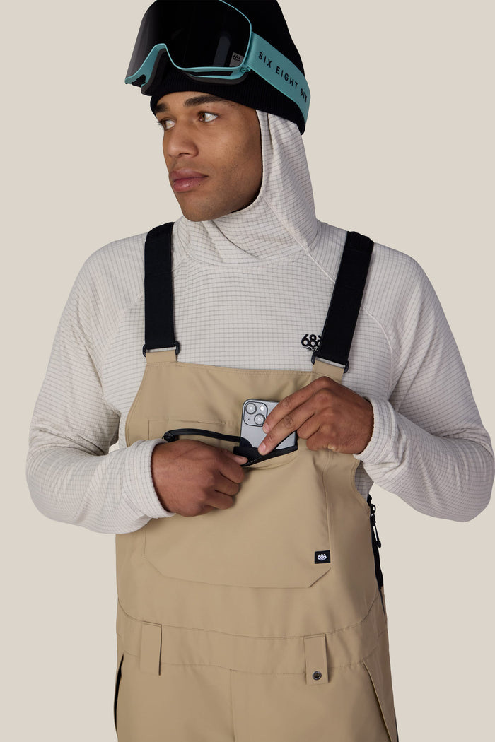 A man wearing the 686 Men’s Lockup™ Shell Bib in beige and a white hoodie, with ski goggles resting on his head, places a smartphone into the bib’s chest pocket while looking to the side against a plain background.