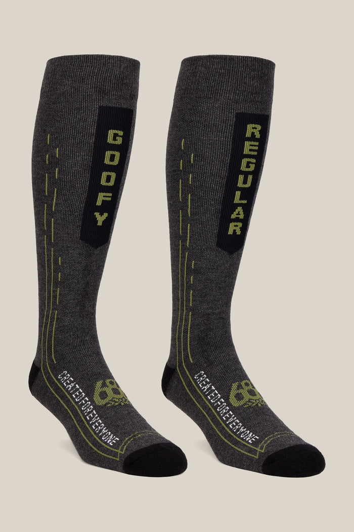 A pair from the 686 Men’s Merino Balance Sock 2-Pack features tall, dark gray performance socks with black toes and heels; one displays “GOOFY” in green text while the other reads “REGULAR.” Both socks include green lines and patterns, subtle 686 branding near the toes, and are designed for optimal comfort and style.
