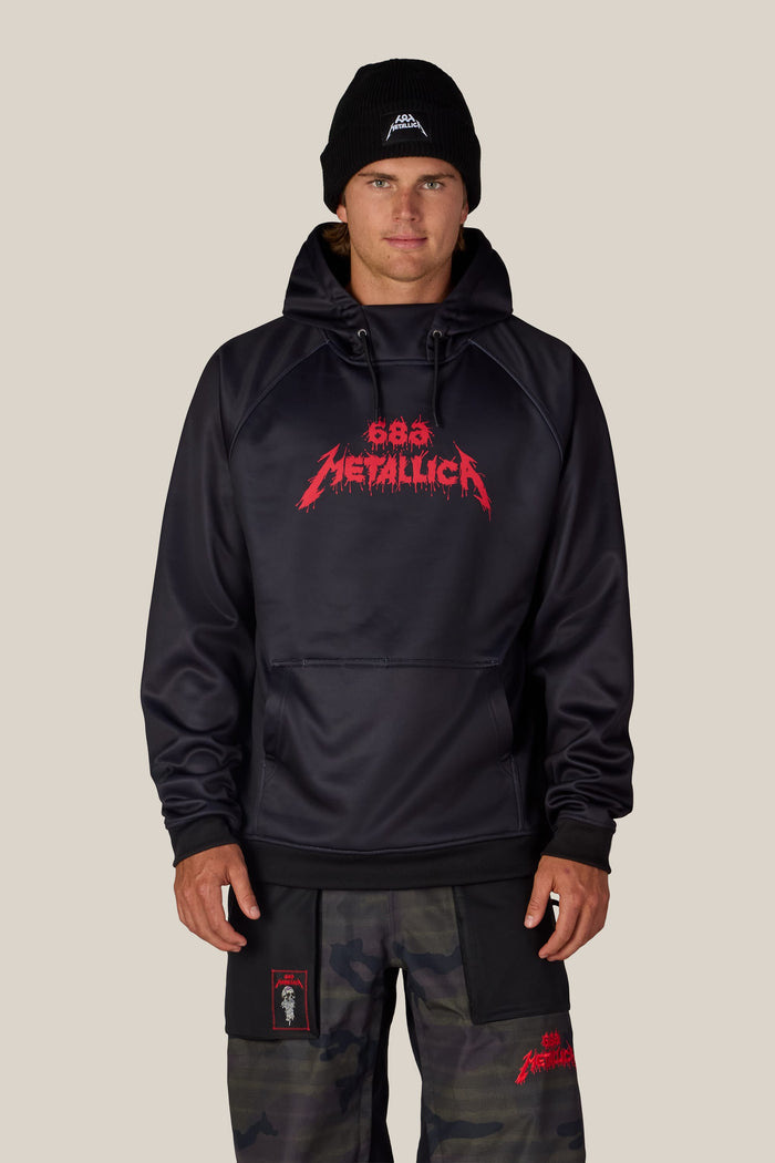 A person wears a black 686 Men’s Metallica Bonded Fleece Pullover Hoody with a red Metallica logo across the chest, a black beanie with a small logo, and camouflage pants.