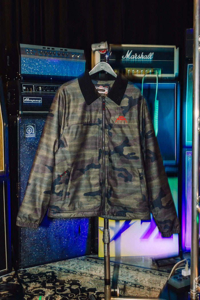 686 Men’s Metallica Caim Jacket in camo-print with a black collar and small red logo on the chest, hanging on a stand in front of music amplifiers.