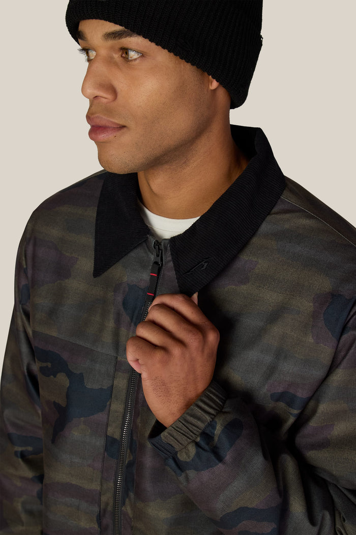 A person wearing a black beanie and a 686 Men’s Metallica Caim Jacket in a camouflage print with a black collar, holding the zipper near the neck.