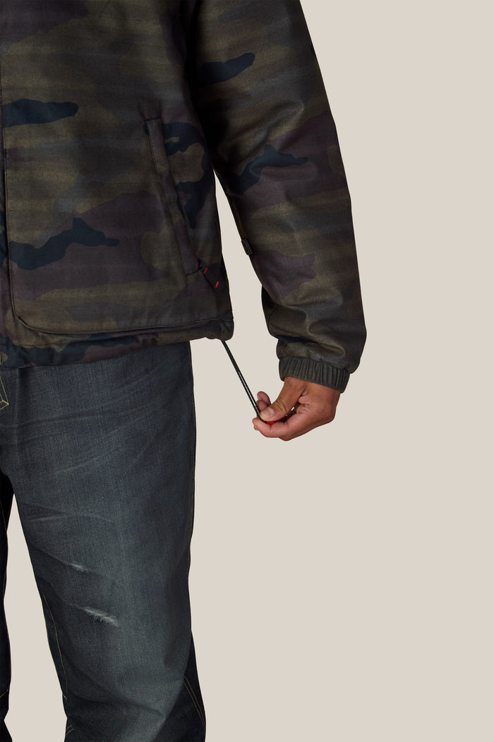 A person wearing a camo-print 686 Men’s Metallica Caim Jacket and dark jeans pulls on the adjustable drawstring at the hem.