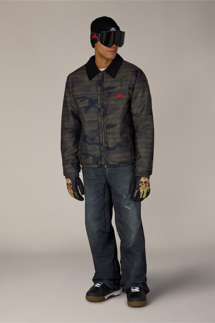 A man wears a 686 Men’s Metallica Caim Jacket with a black collar, dark jeans, a black beanie, ski goggles, and gloves featuring skeleton graphics.