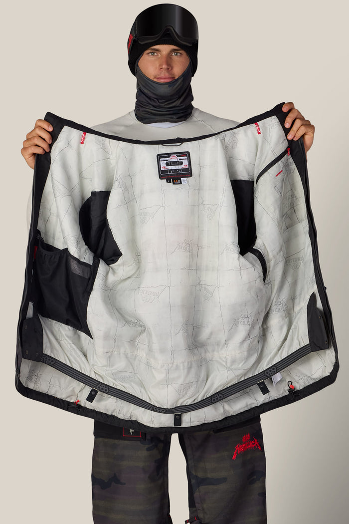 A person in a ski mask and goggles holds open a black 686 Men’s Metallica One Insulated Jacket, revealing a white interior lining patterned with Metallica logos and a cassette tape graphic.