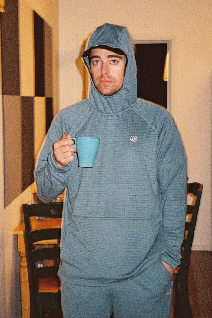 A man wearing a blue 686 Men’s Mission Grid™ Raglan Hoody stands indoors, holding a blue mug and looking at the camera with a neutral expression. Chairs and a table are visible in the background.