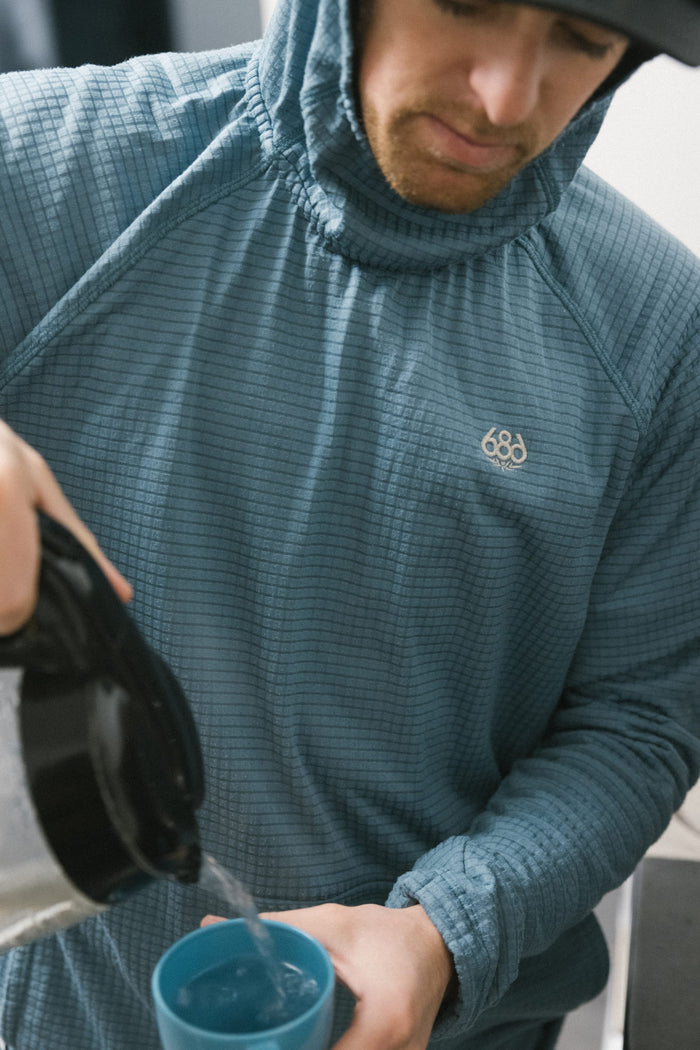 Wearing the 686 Men’s Mission Grid™ Raglan Hoody by 686, a person pours hot water from a black kettle into a blue mug, with their face partially visible and head slightly tilted down.