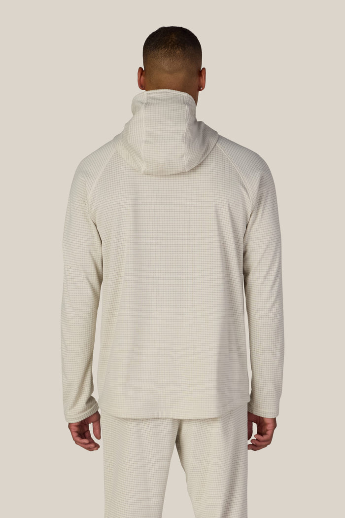 A person stands facing away, wearing the 686 Men’s Mission Grid™ Raglan Hoody in a light color with matching pants. The outfit showcases a subtle checkered pattern, enhancing its contemporary style against a plain, neutral background.