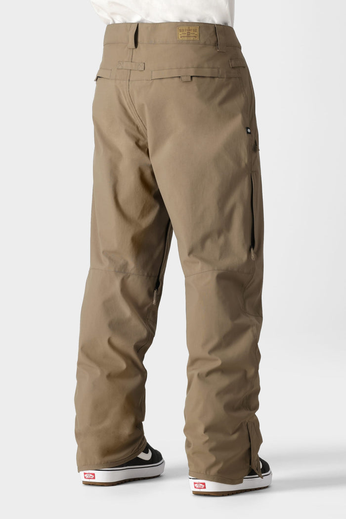A person wearing the 686 Men’s Standard Shell Pant by 686—loose-fitting, affordable khaki pants with pockets—paired with black-and-white Vans sneakers, stands against a plain light background. The back view highlights seam and waistband details on the pants.