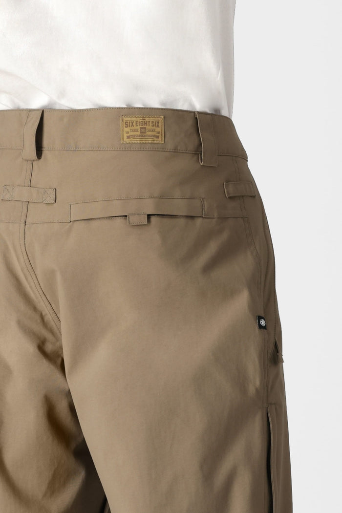 Close-up of a person wearing the 686 Men’s Standard Shell Pant in tan with a white shirt, highlighting belt loops, rear pocket details, and a rectangular brown 686 logo patch above the right pocket. These budget-friendly shell pants from 686 are crafted from waterproof material for enhanced functionality.