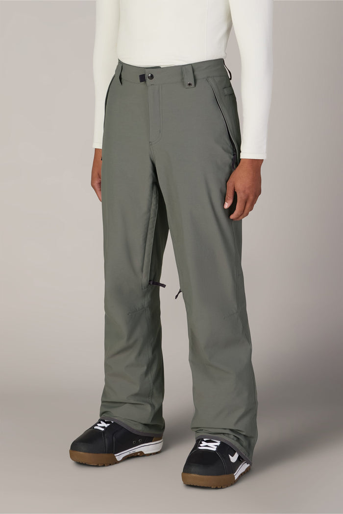 A person wearing 686 Men’s Standard Shell Pant in sage green, a white long-sleeve top, and black and white snow boots stands against a plain light gray background.
