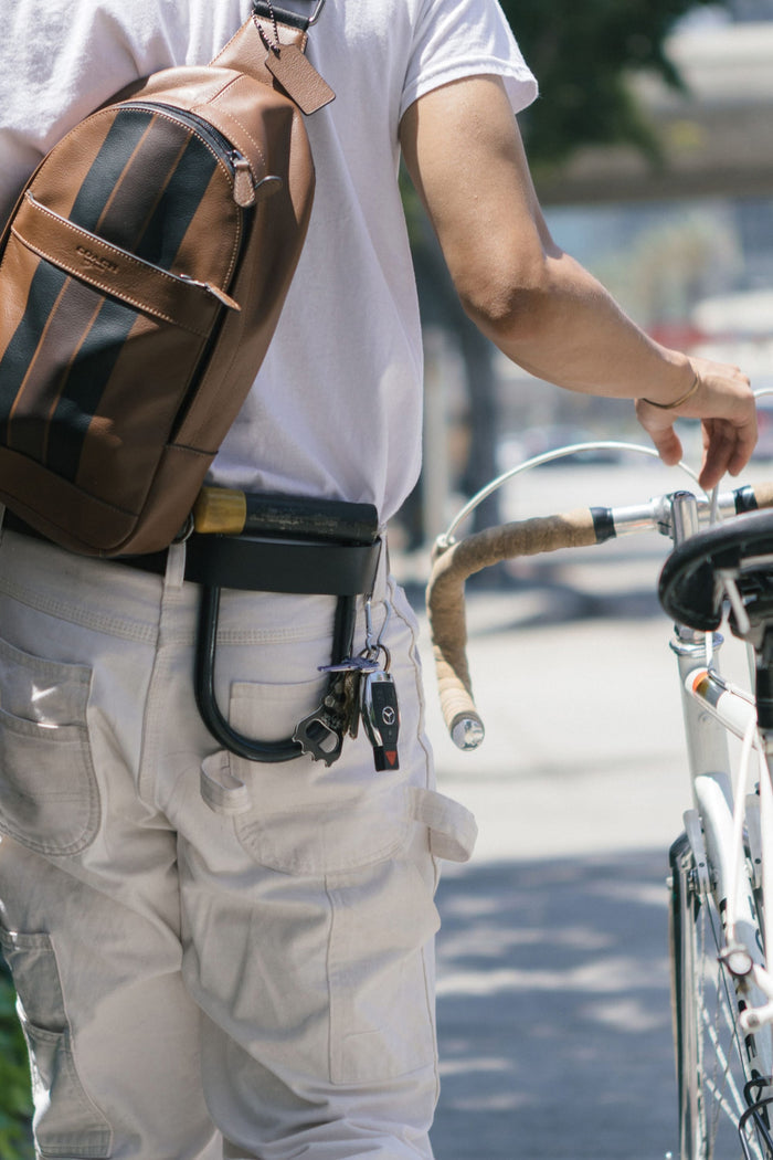 A person in white pants and shirt walks a bicycle outdoors on a sunny day, carrying a brown backpack. Keys and the Westwell 686 Westwell Voyager Belt bike lock hang from their belt.