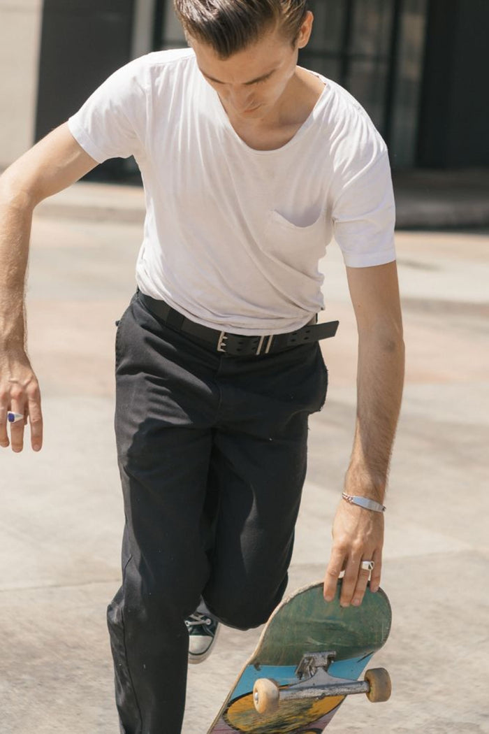 Wearing a white t-shirt, black pants, and a 686 Westwell Voyager Belt by Westwell, a person is about to step onto a skateboard outdoors‚Äîone hand reaching forward, the other holding their skateboard.