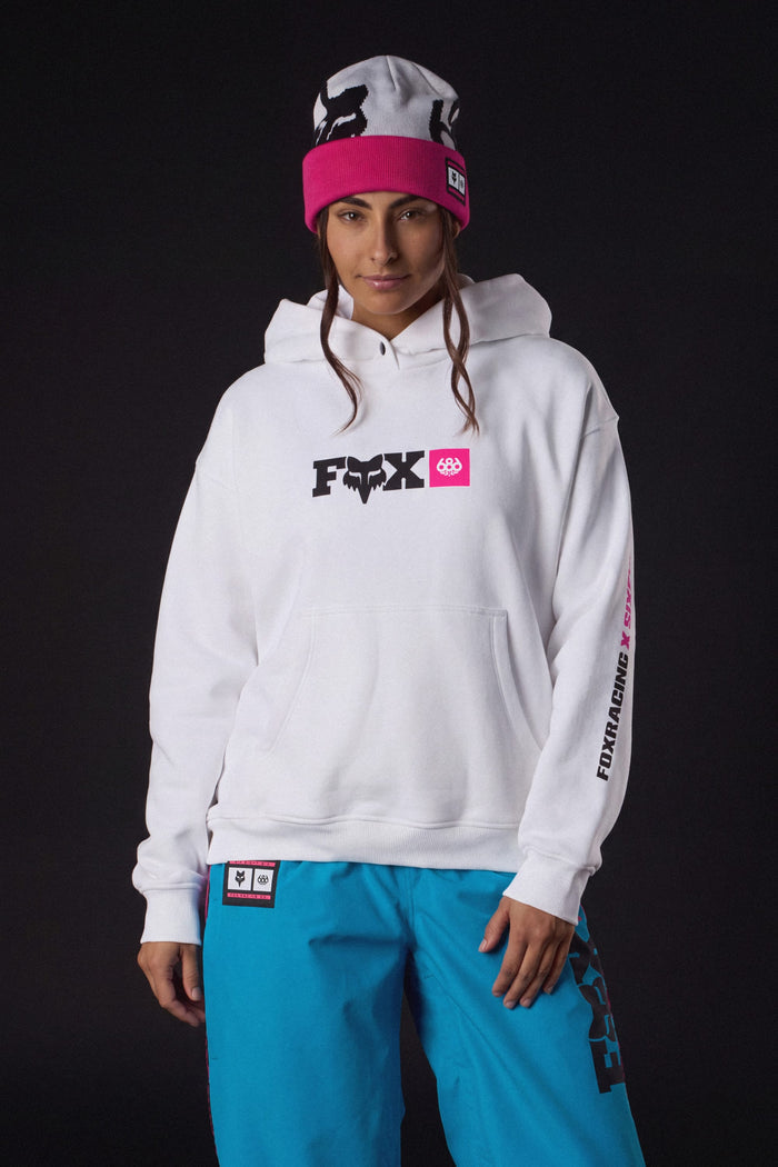A person wearing a 686 Women’s Fox Racing Premium Heavyweight Pullover Hoody, a pink and white knit beanie, and bright blue pants.