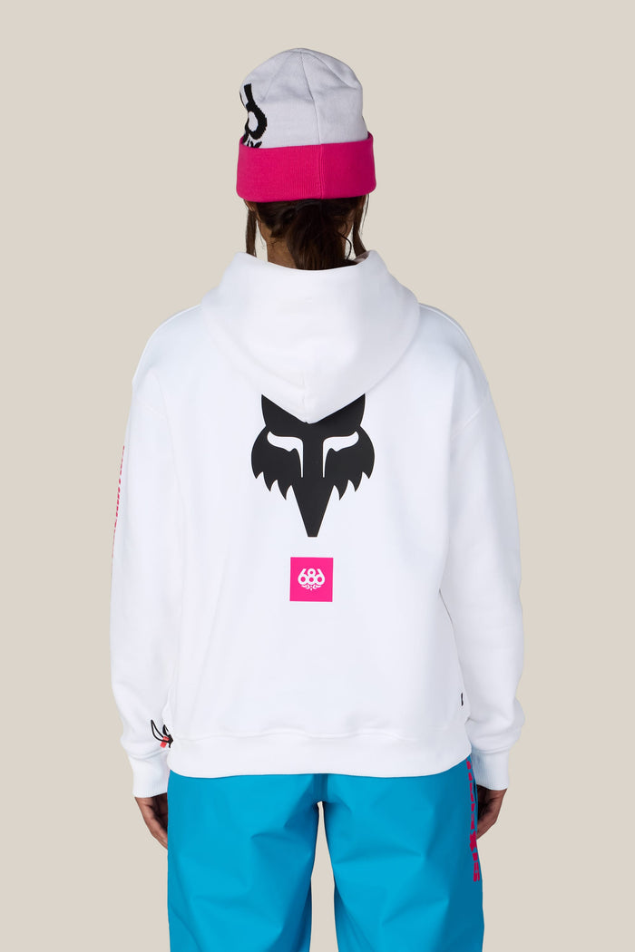 A person seen from behind wears a 686 Women’s Fox Racing Premium Heavyweight Pullover Hoody paired with a pink and grey beanie and bright blue pants.