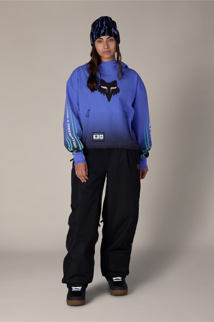 A person wearing a 686 Women’s Fox Racing Waterproof Crop Hoody, black snow pants, and a patterned beanie.