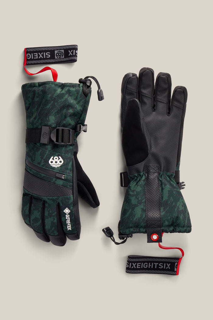 A pair of 686 Women’s GORE-TEX® SMARTY® 3-In-1 Gauntlet Gloves featuring green camouflage with black palms and adjustable straps. Red and black 686-branded wrist leashes are attached. The gloves include a textured grip and drawcords at the cuffs.