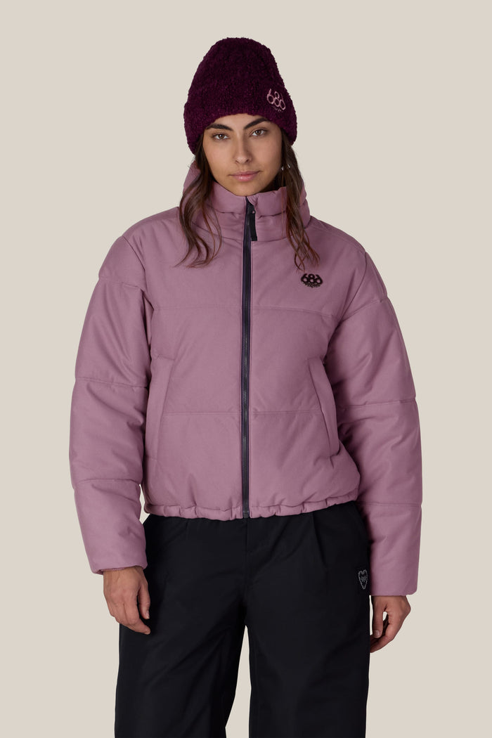 A person wearing the 686 Women’s Gracious Puffer Jacket in pink, paired with black pants and a purple knit beanie, stands facing the camera against a plain light background.