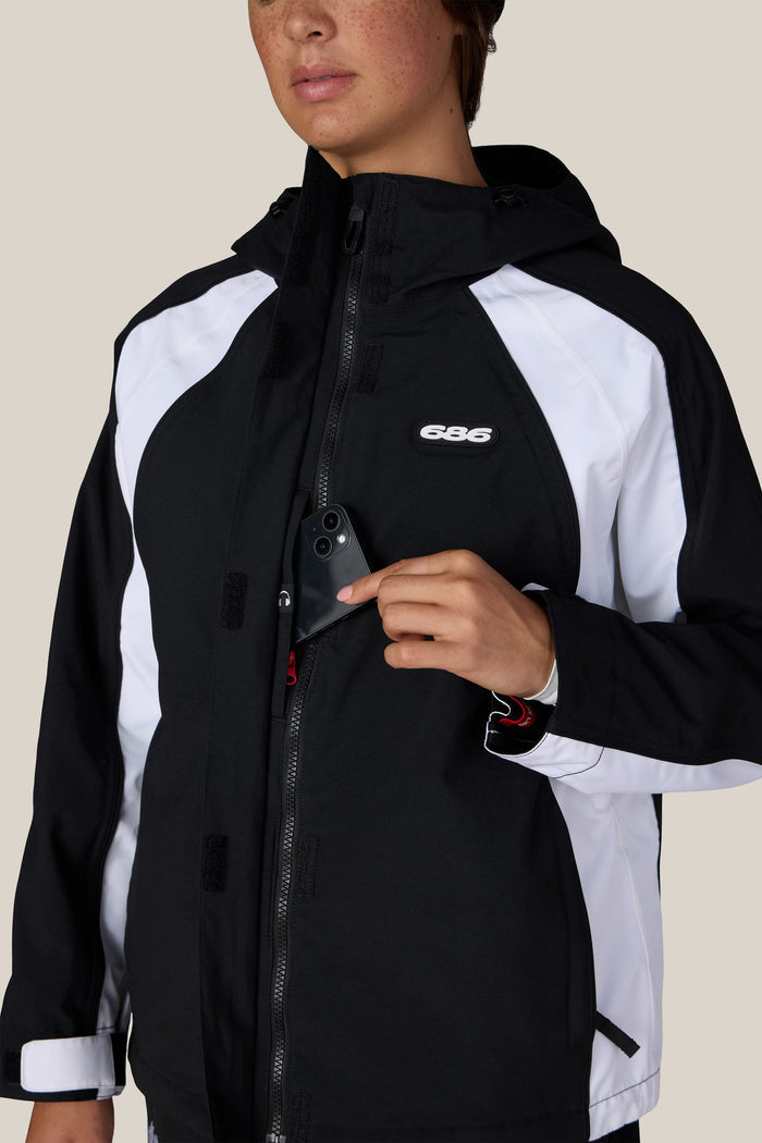 A person wearing the 686 Women’s Hologram™ Shell Jacket in black and white, featuring visible zippers, Velcro, and the 686 logo, places a phone into the chest pocket. The plain beige background contrasts with their partially visible face.