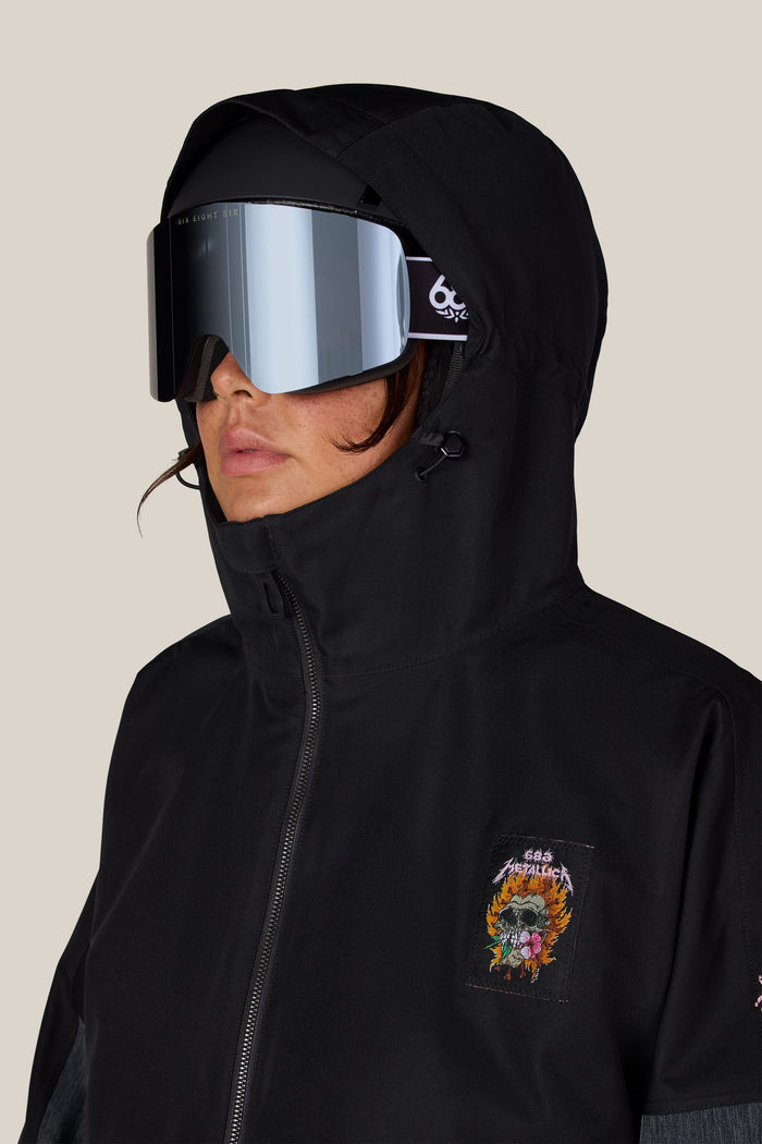 A person wearing a black 686 Women’s Metallica Burning Skull Insulated Jacket with a hood, black helmet, mirrored ski goggles, and a flaming skull patch on the chest.
